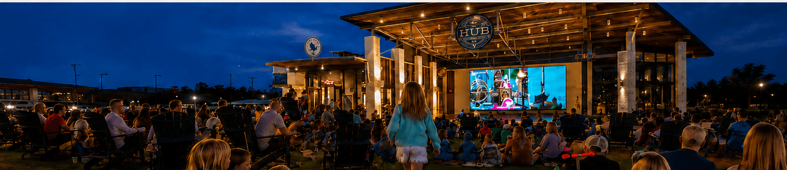 Families gathered at an outdoor movie night at The HUB, families seated on a lawn watching a large LED screen at blue hour.